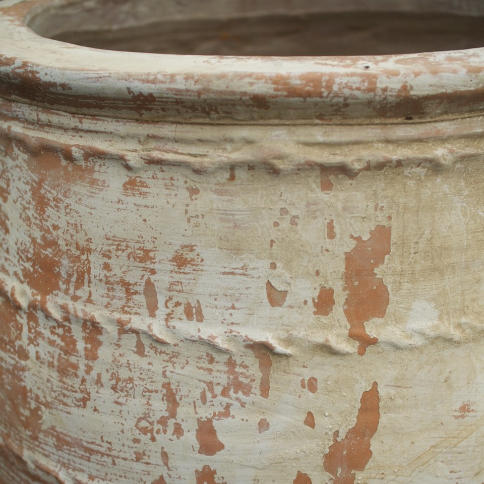 Close-up of a large rustic ceramic planter pot with a distressed finish, showing a cream-colored glaze with terracotta brown patches and horizontal ridges.
