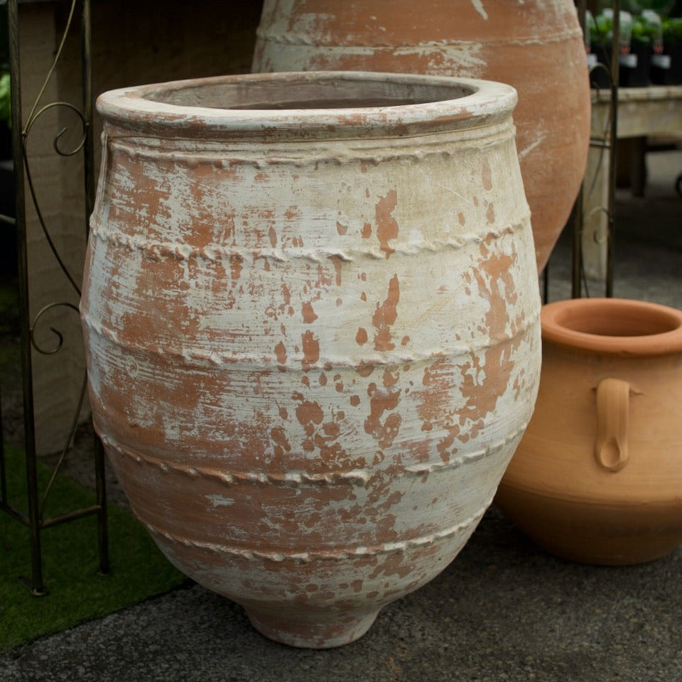 A large distressed terracotta planter pot with a weathered off-white and brown finish, shown alongside other rustic clay pots in an outdoor garden setting.