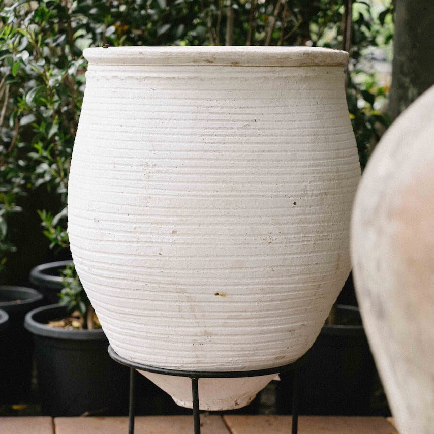 A large ribbed off-white ceramic planter pot displayed on a black metal stand, set on a tiled patio with green foliage in the background.