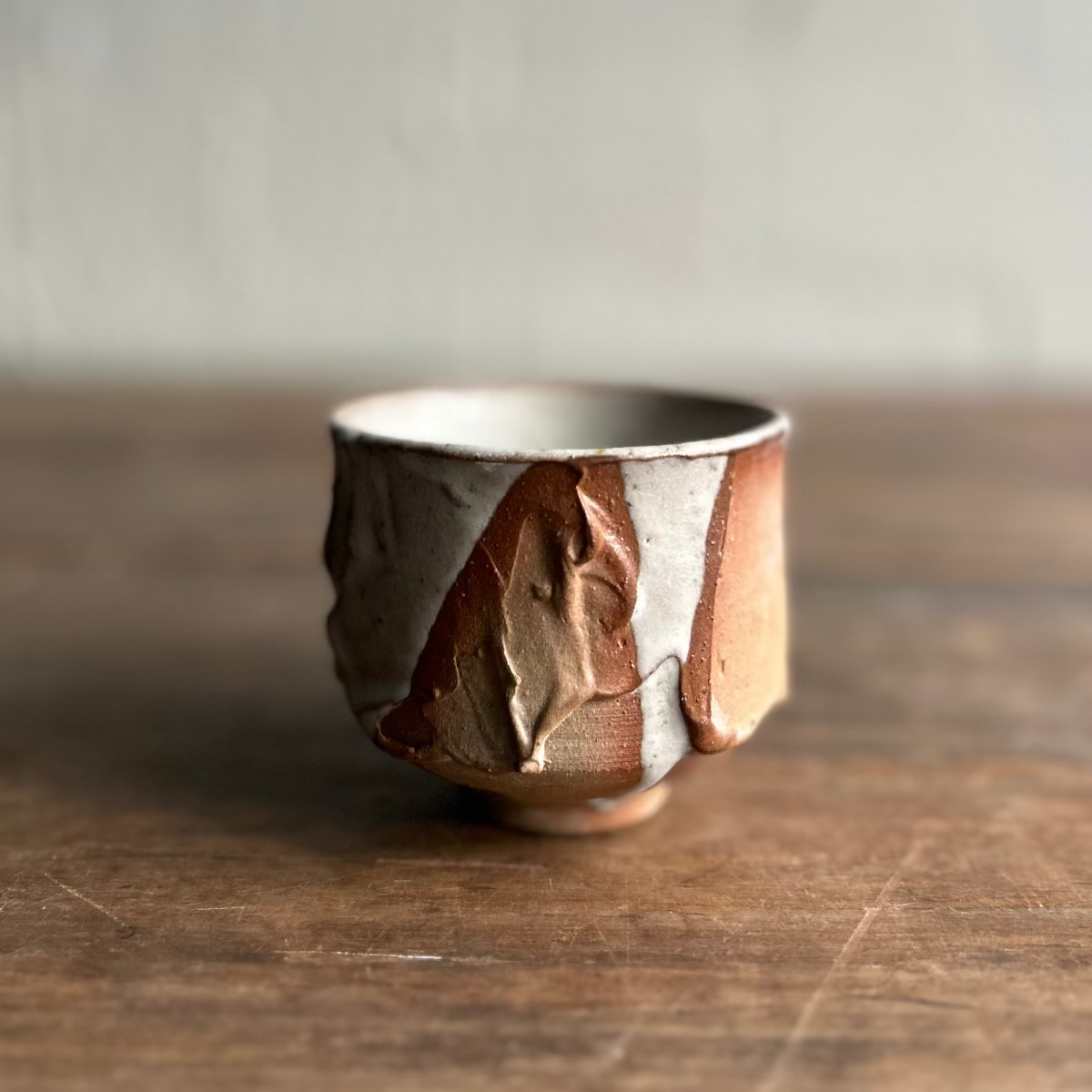 A handmade ceramic tea cup with a textured, organic ridged glaze in earthy brown, beige, and white tones, sitting on a rustic wooden table against a neutral gray wall.
