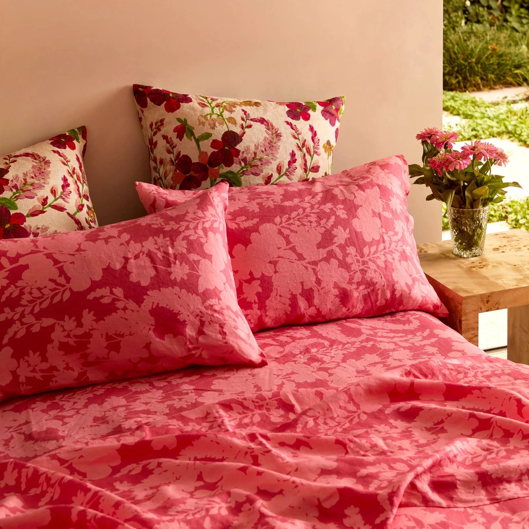 Pink floral bedding set with matching pillows on a bed, next to a wooden side table with flowers.