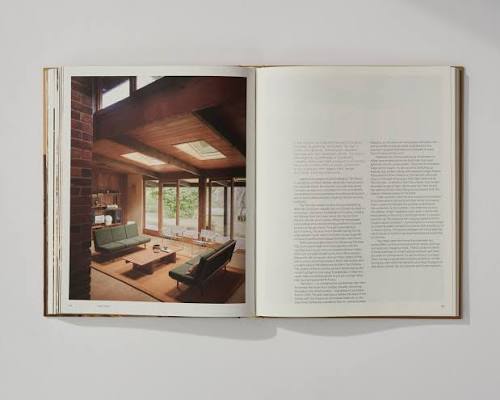 An open architectural book or magazine spread, showing a photograph of a modern living room with large windows and wood ceiling on the left page, and columns of text on the right page, photographed against a light gray background.