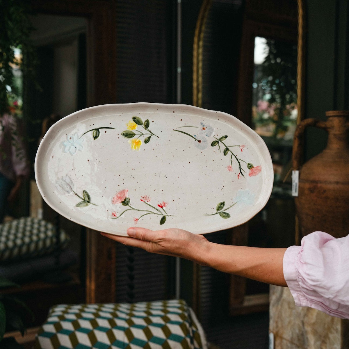 A hand holds a large, oval, off-white ceramic serving platter decorated with hand-painted green vines, pink and yellow flowers, and subtle speckles. The background shows a rustic, vintage-style interior with wooden furniture and mirrors.