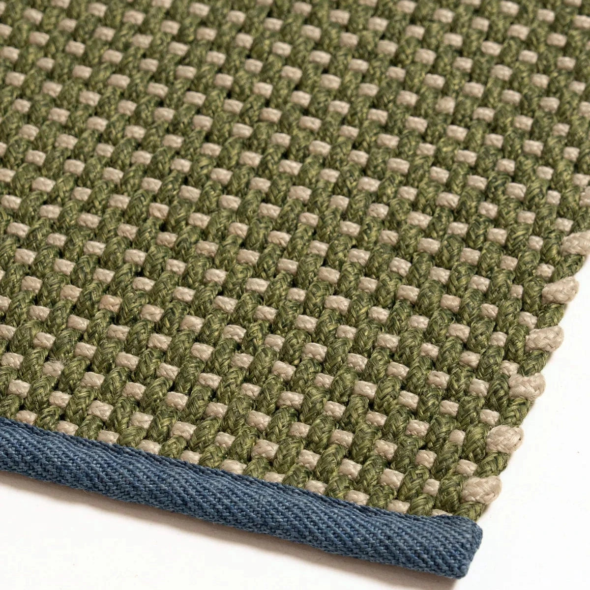 Close-up of a woven rug corner showing an olive green and beige basketweave pattern with a blue-gray twisted edge.
