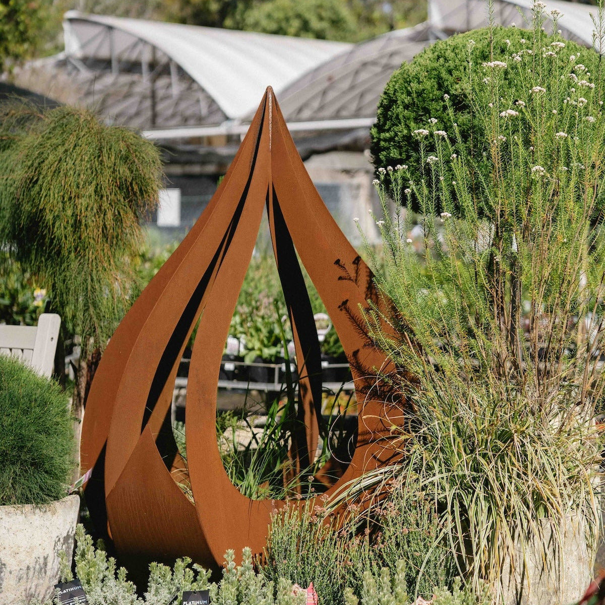 A rusted corrugated metal garden sculpture shaped like a stylized teardrop or abstract leaf, displayed in a landscaped garden bed with plants and a greenhouse in the background.