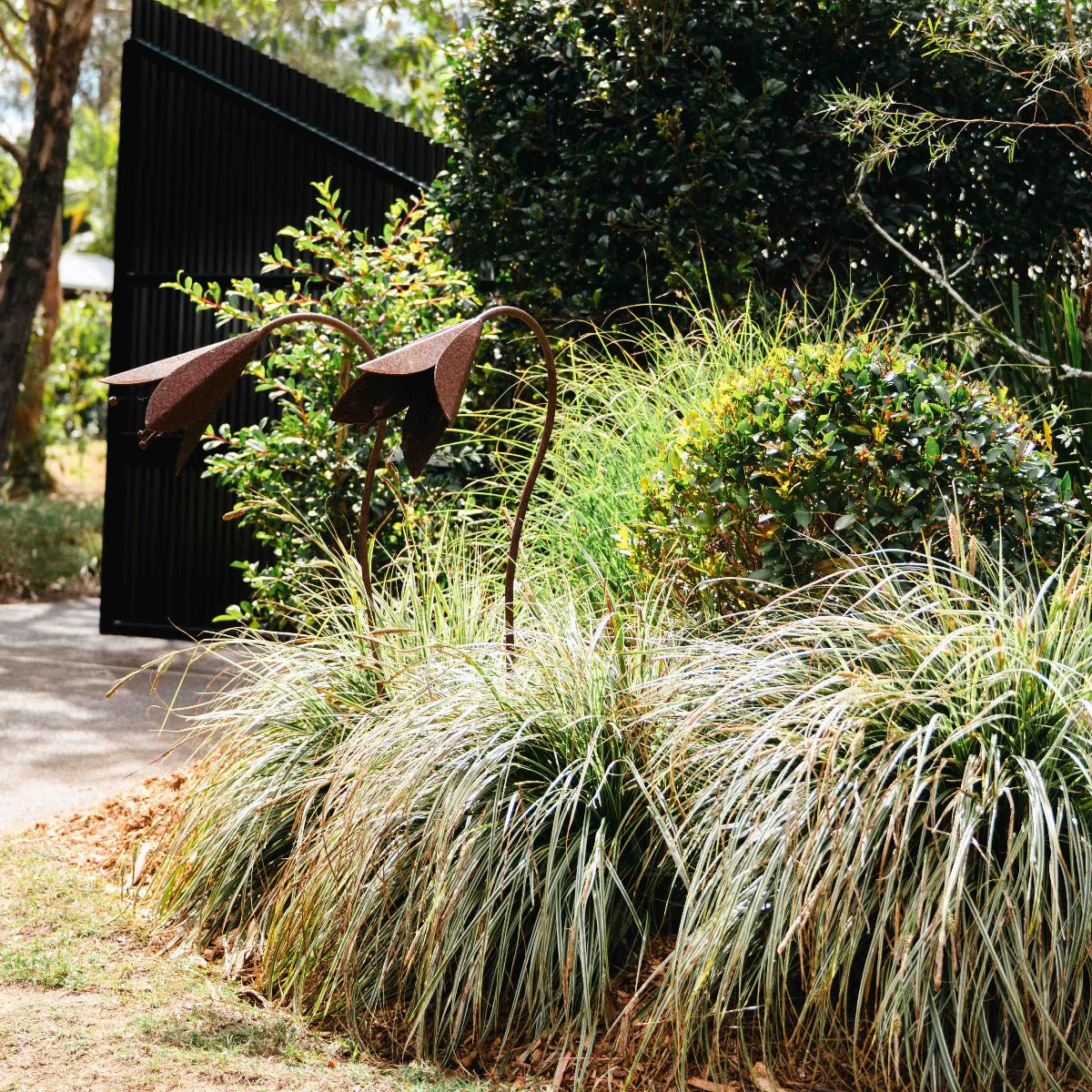 A landscaped garden bed with tall ornamental grasses, dense shrubs, and a dark vertical slatted wall behind them. Two sculptural metal garden lights with curved arms and leaf-like shades are placed among the plants beside a light-colored path.