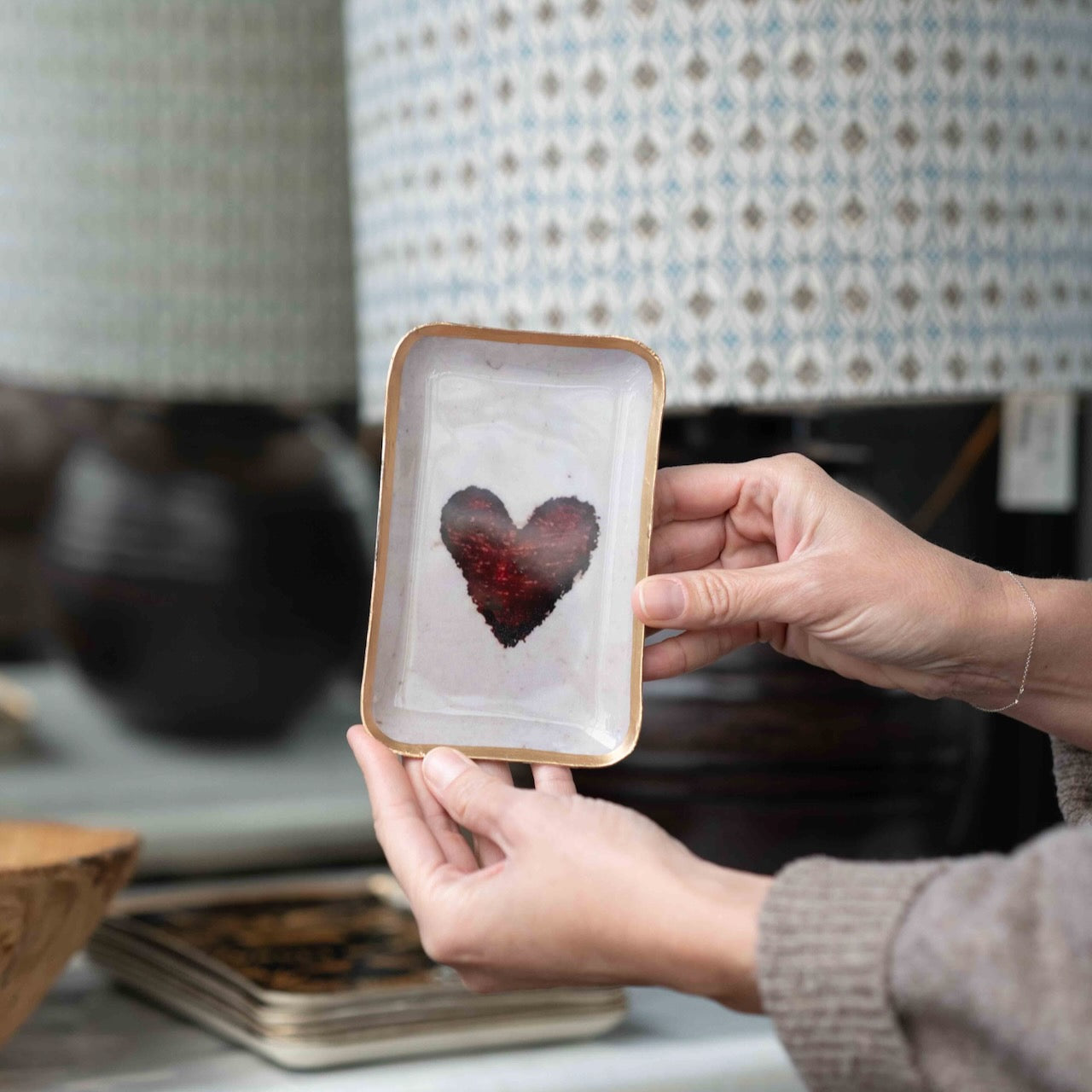 A person holds a small rectangular ceramic plate with a gold-toned rim and a textured red heart design in the center, set against a softly blurred home background.
