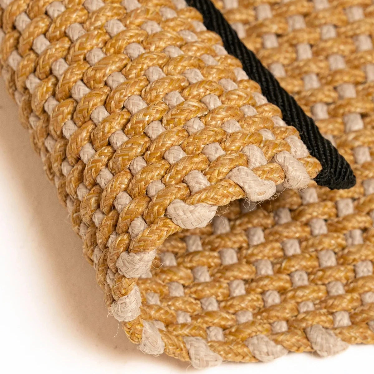 A close-up of a woven mat or rug with a textured pattern in mustard yellow and light beige, rolled over to show the underside and a black edge trim, on a white background.