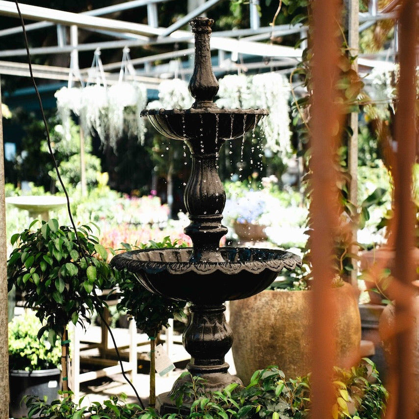 A dark metal tiered garden fountain with cascading water, set in a lush greenhouse or garden with green plants and a metal trellis background.