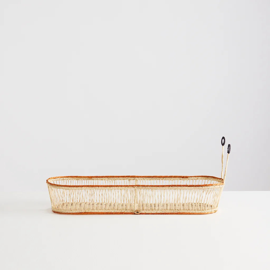 A long, narrow woven basket with a natural beige body and a thin orange-brown rim, featuring two small black hanging tags on the right side, photographed on a white surface against a white background.