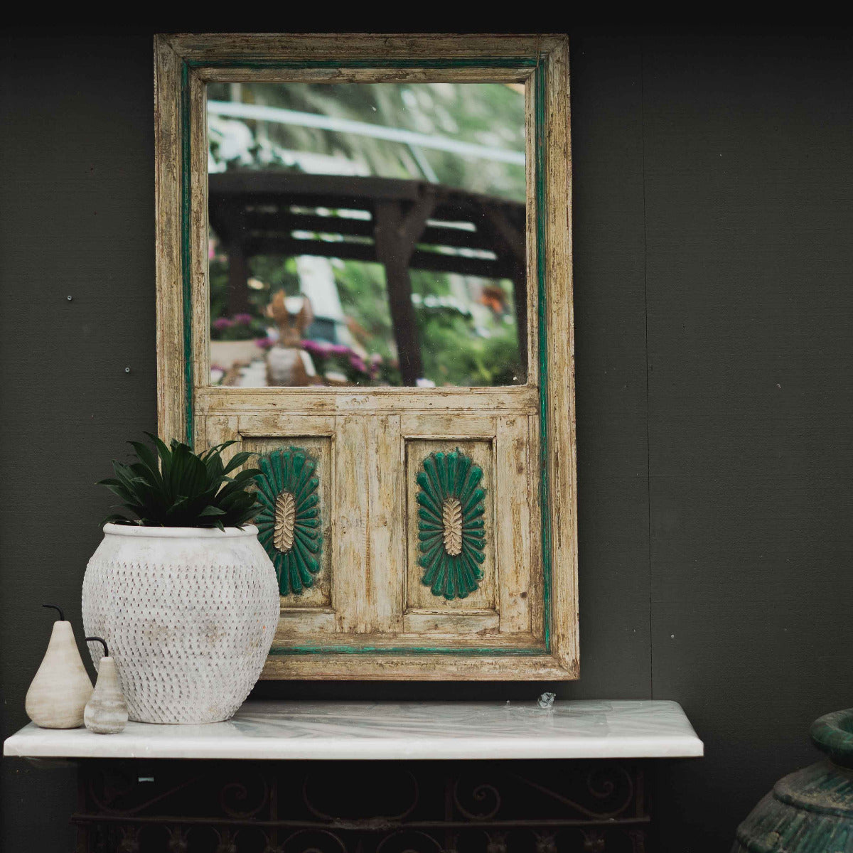 A rustic, distressed wooden wall mirror with a green painted frame and decorative leaf motifs, displayed on a white marble console table against a dark gray wall.