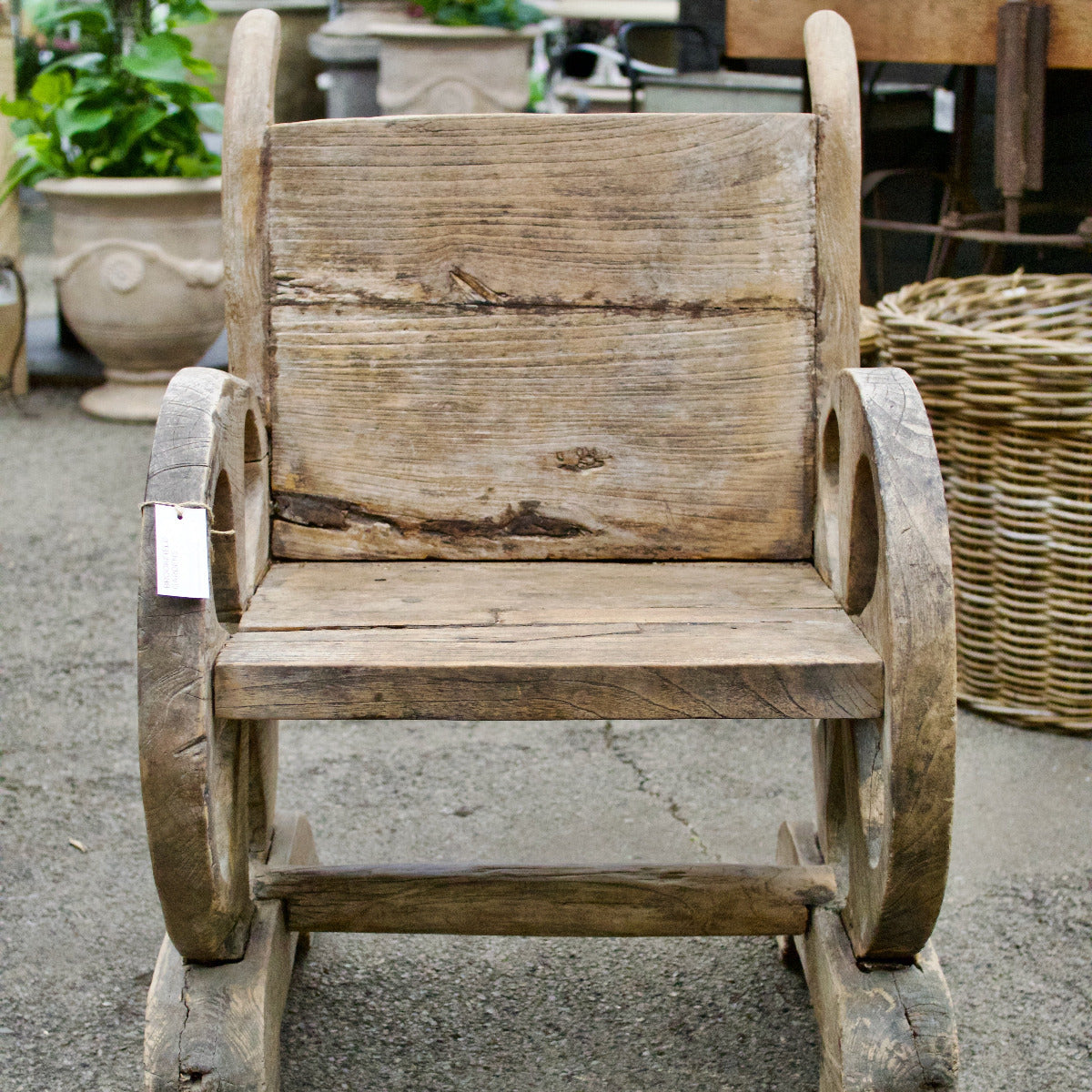 Wooden wheelbarrow-style chair on a concrete surface with plants and baskets in the background