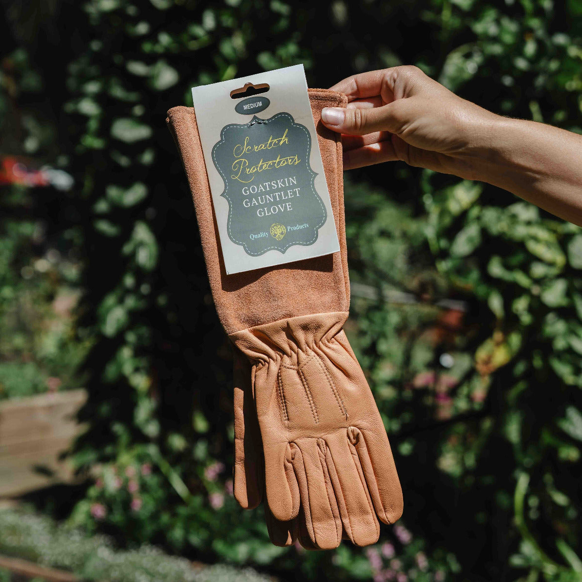 Pair of brown leather gloves with a label held by a hand against a green foliage background