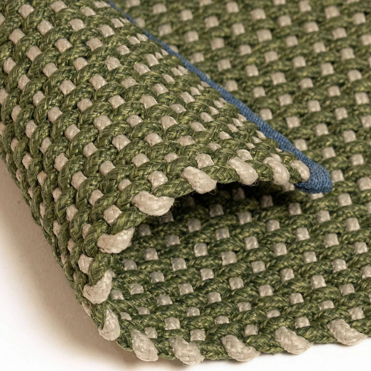 A close-up of a woven outdoor rug with a textured green and beige basketweave pattern, folded over to show its underside and a blue backing layer.