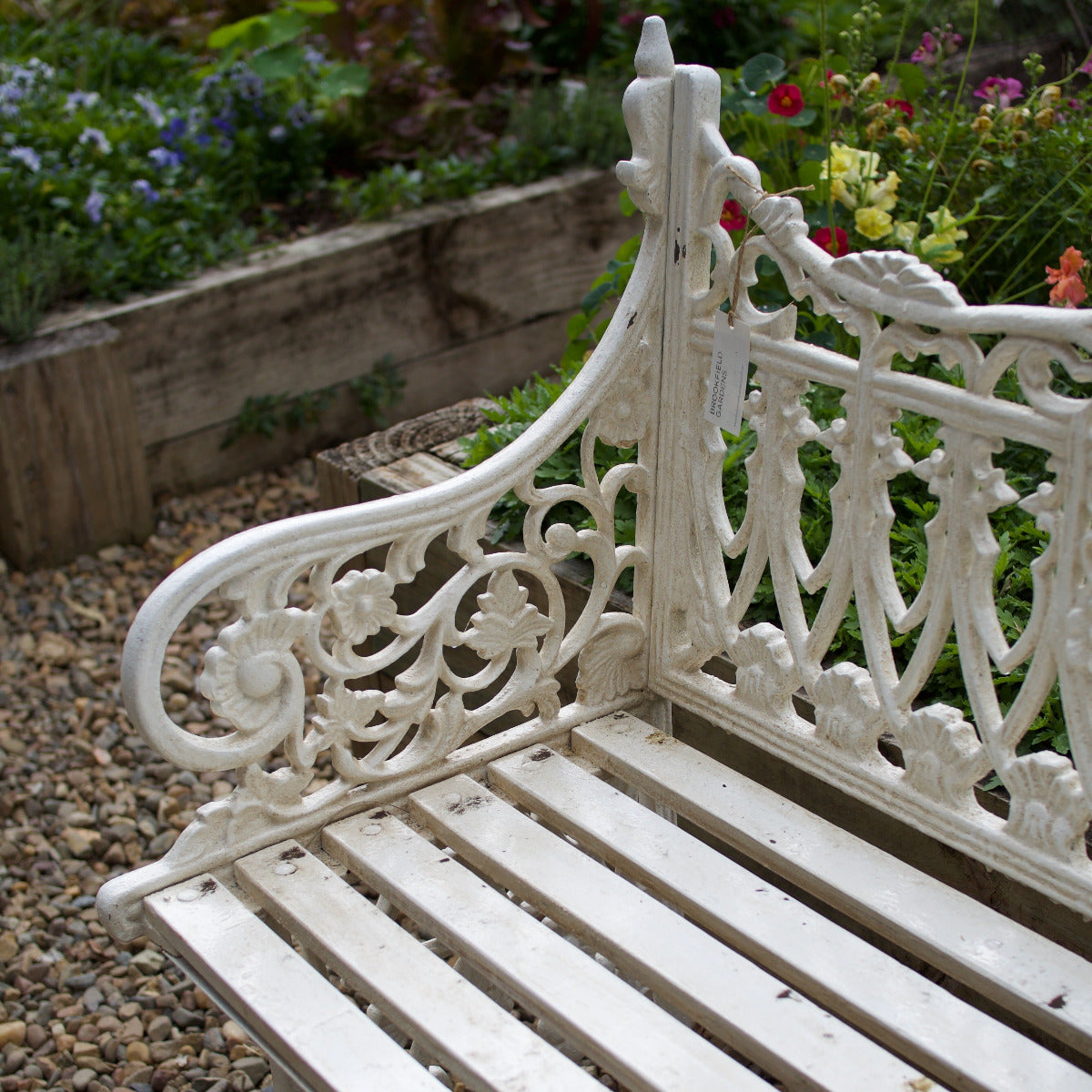 A white cast iron garden bench with ornate scrollwork and slatted seat, photographed outdoors in a garden with gravel and flowers in the background.