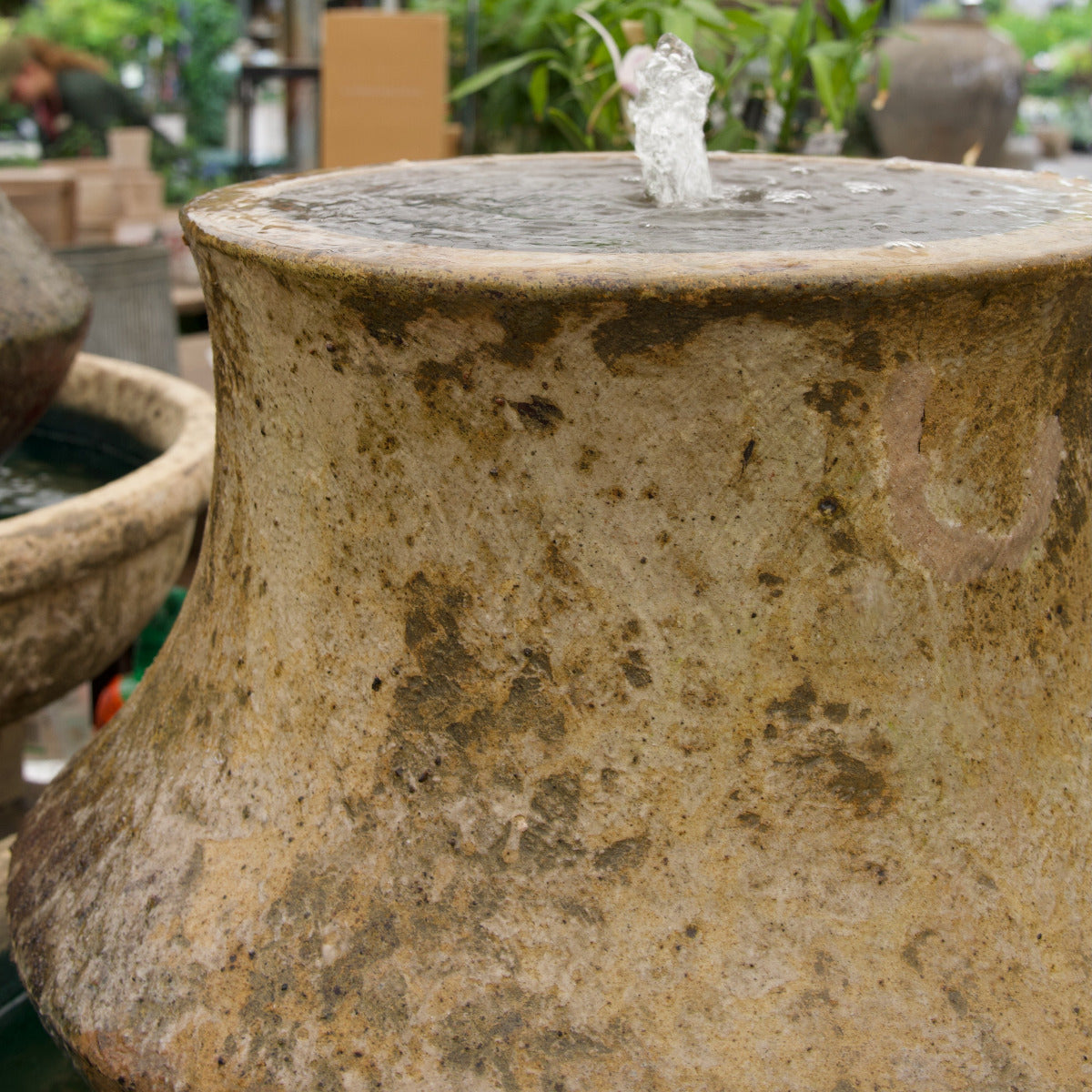 Vintage stone vase with water spilling out, surrounded by greenery and other planters.