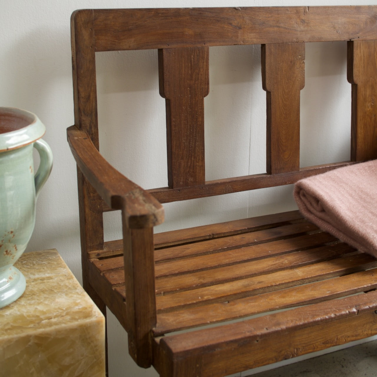 A close-up of a rustic wooden bench with a warm medium-brown finish, featuring a slatted seat and backrest. A folded dusty pink blanket is draped over the right side, and a light green ceramic vase sits on a marble side table to the left against a plain off-white wall.