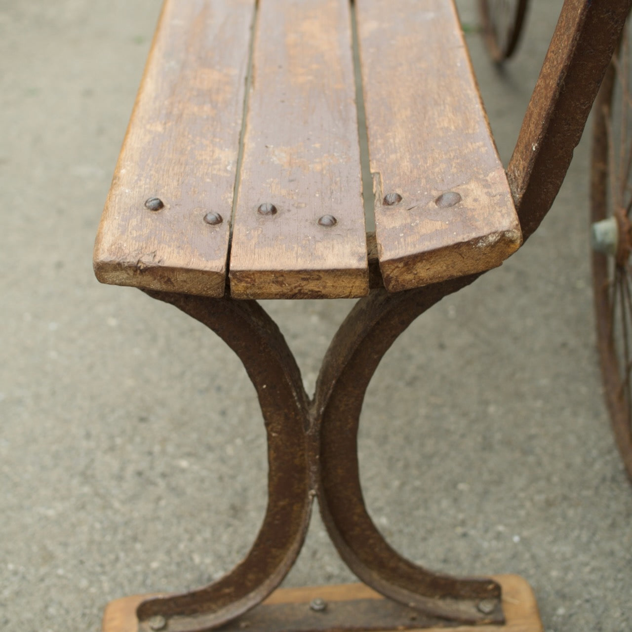 Close-up of a rustic wooden bench with weathered brown slats and a rusted metal frame, photographed outdoors on a gray concrete surface.