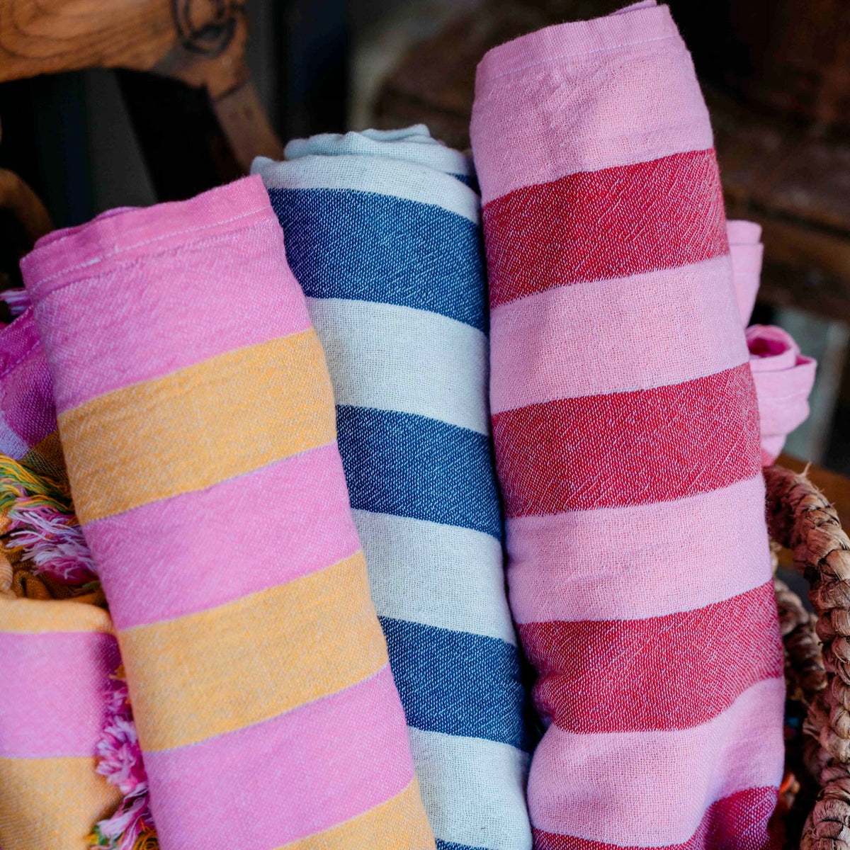 Three rolled striped textiles, likely towels or blankets, displayed in a basket. The left and right rolls feature pink and yellow stripes, while the center roll has blue and white stripes.