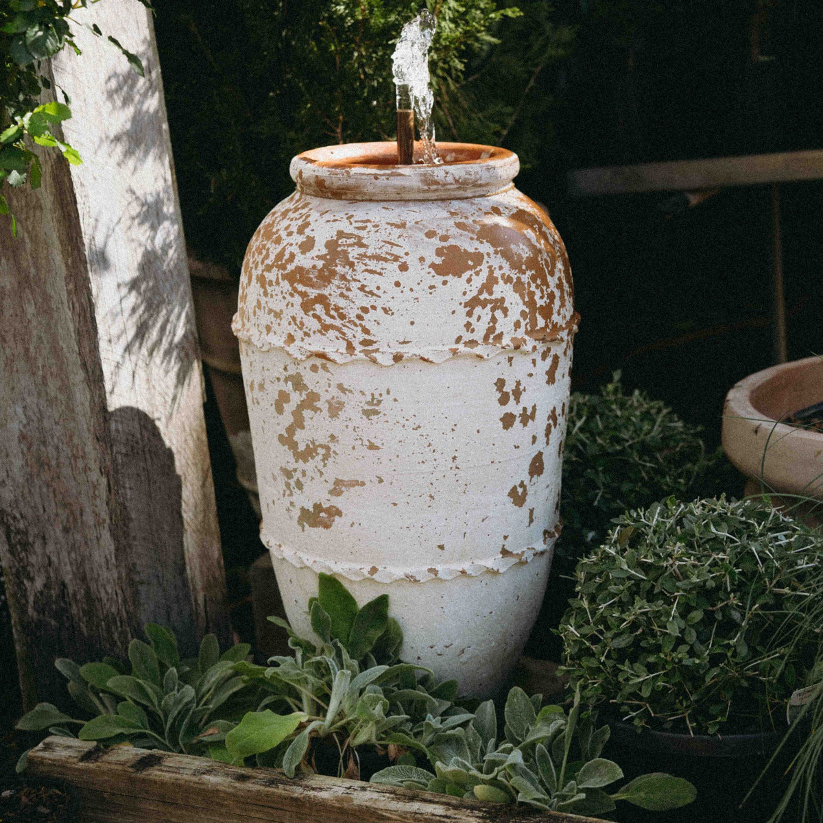 Weathered Turkish Odemis pot fountain with a simple copper water spout surrounded by garden plants.