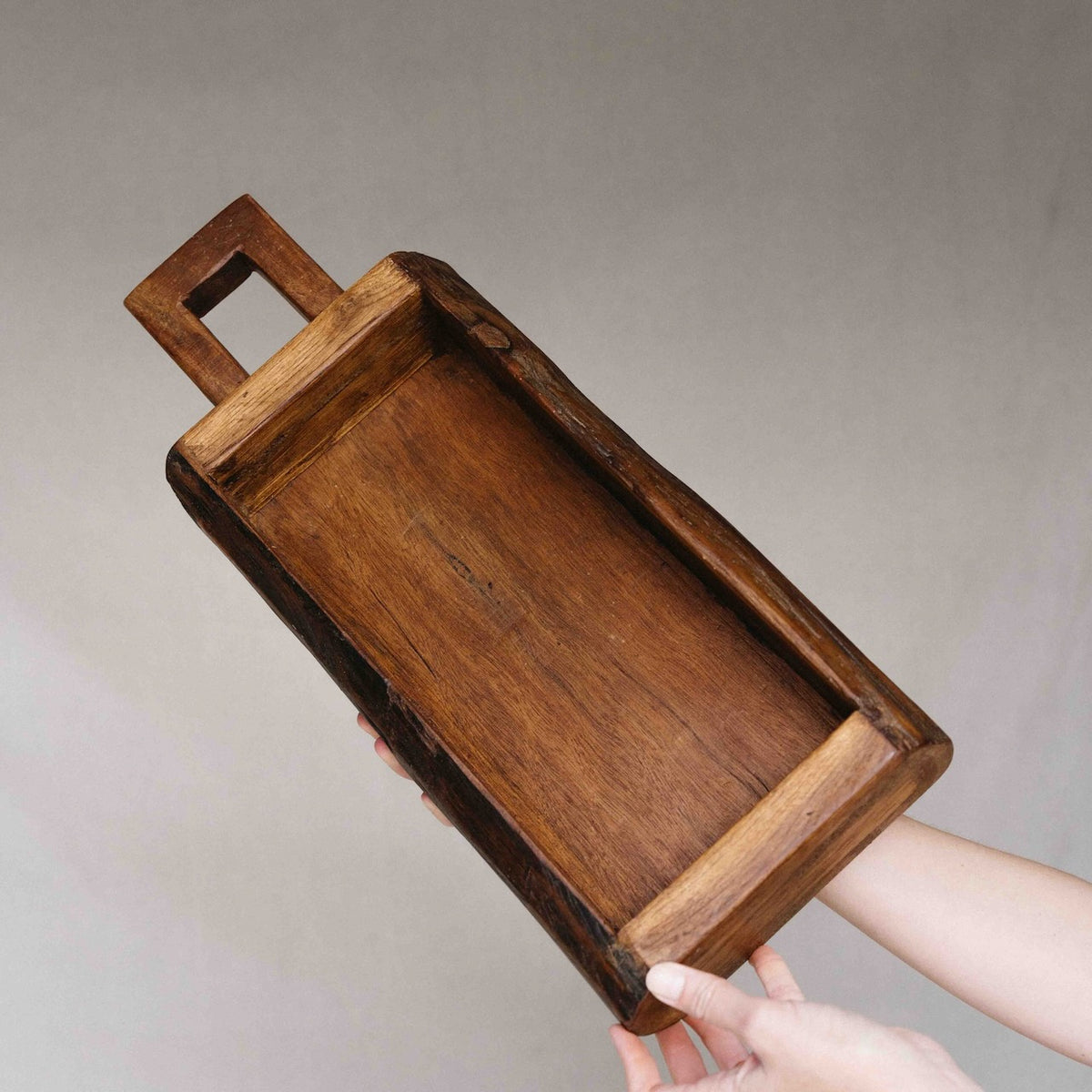 A rectangular dark brown wooden serving tray with a cutout handle, held by two hands against a neutral gray background.