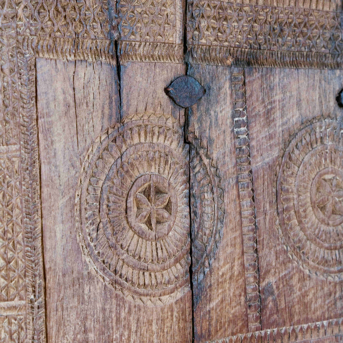 Close-up of an intricately carved wooden door or panel with circular geometric and floral engravings, showing natural wood grain and a dark metal latch near the top.