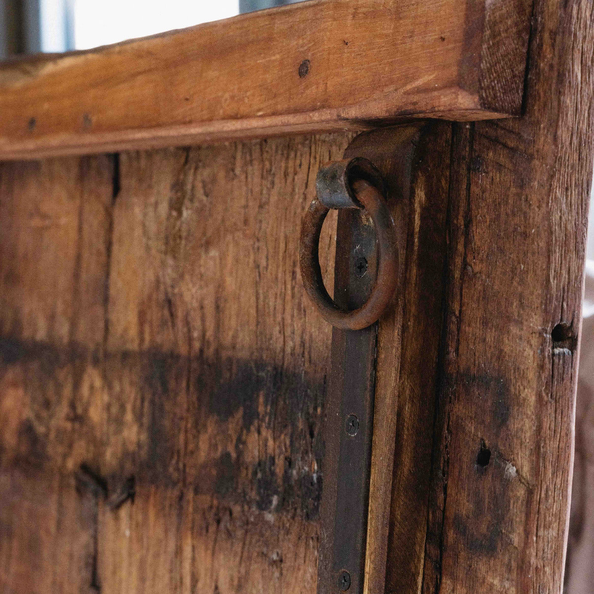 Close-up of a rustic wooden door with a dark metal hinge and a round ring pull handle, showing weathered wood grain and aged metal details.