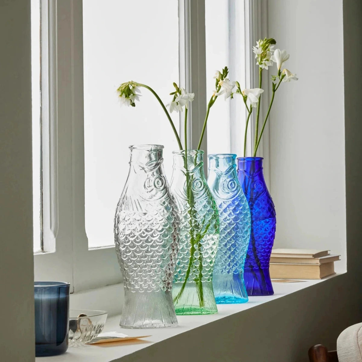 Three glass vases of different colors on a windowsill with flowers.