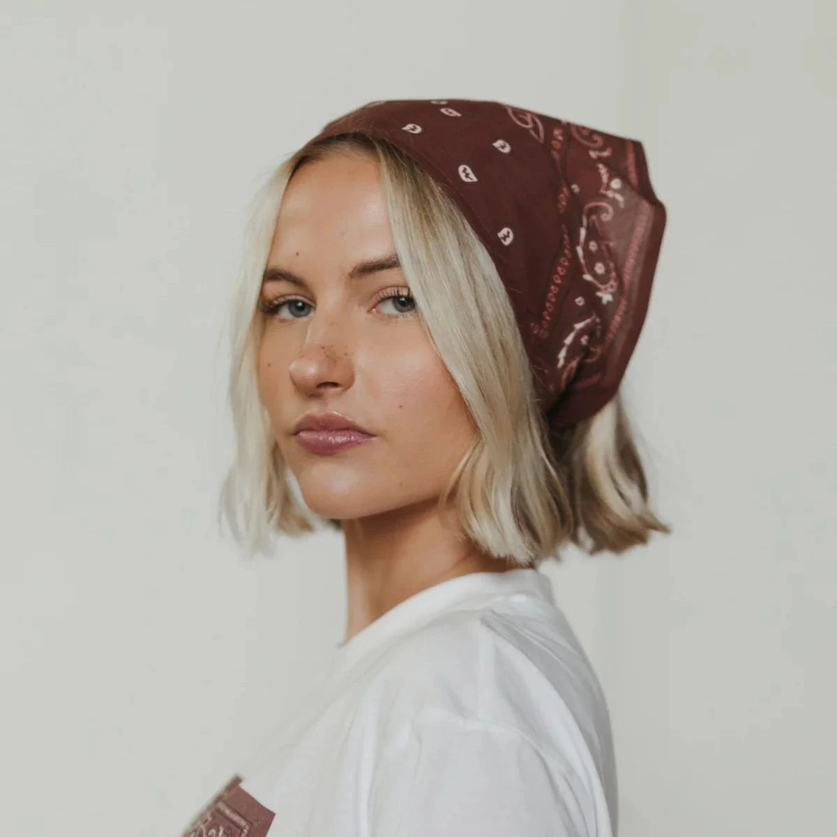 A woman with short blonde hair wearing a brown bandana with a white paisley pattern, styled as a headscarf, paired with a white t-shirt against a plain light background.