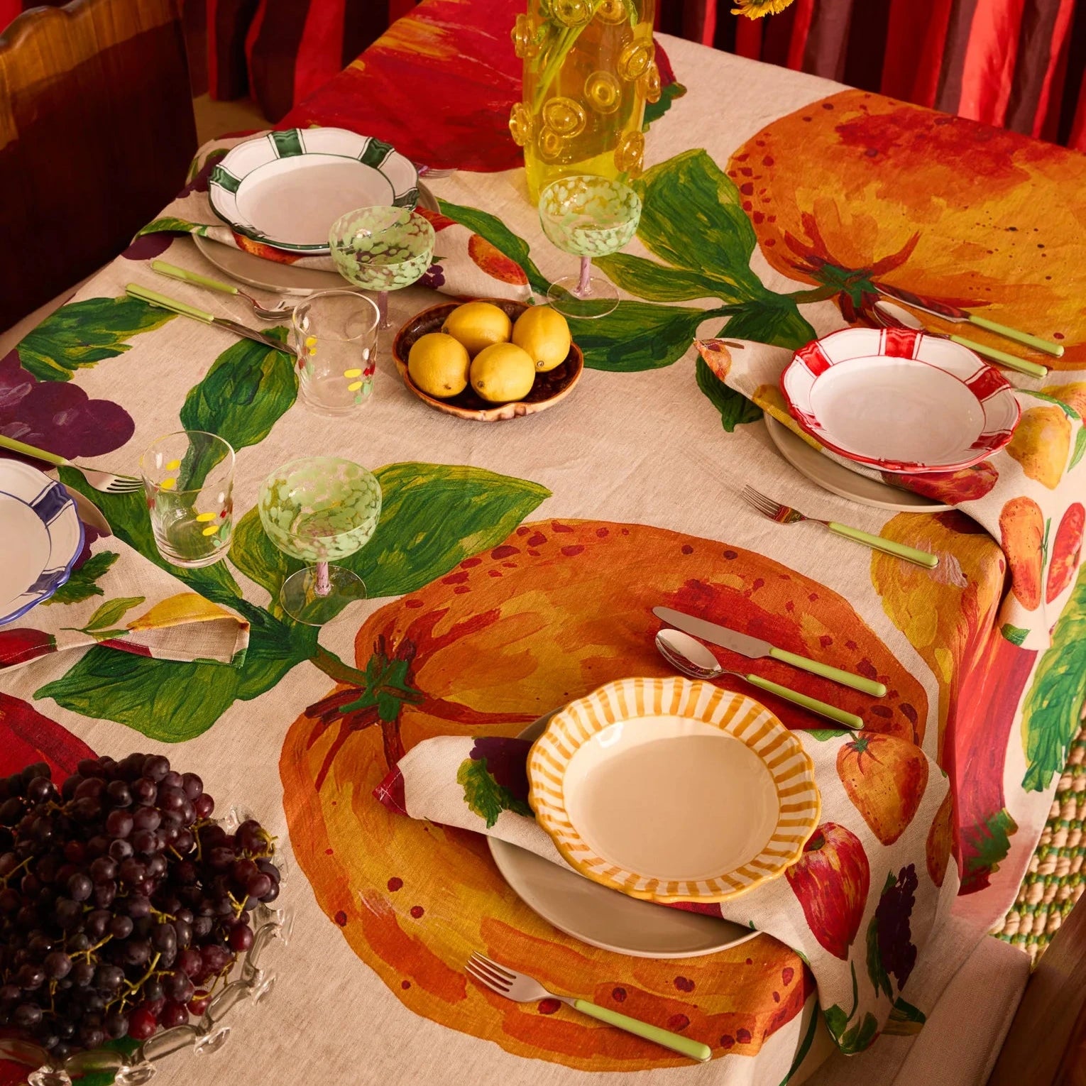 Decorative table setting with a colorful tablecloth featuring fruits and vegetables, surrounded by red curtains.