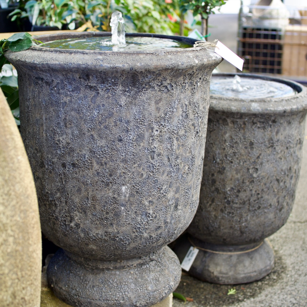 Two textured stone urn-style outdoor water fountains with small clear water jets, shown in a garden or patio setting.
