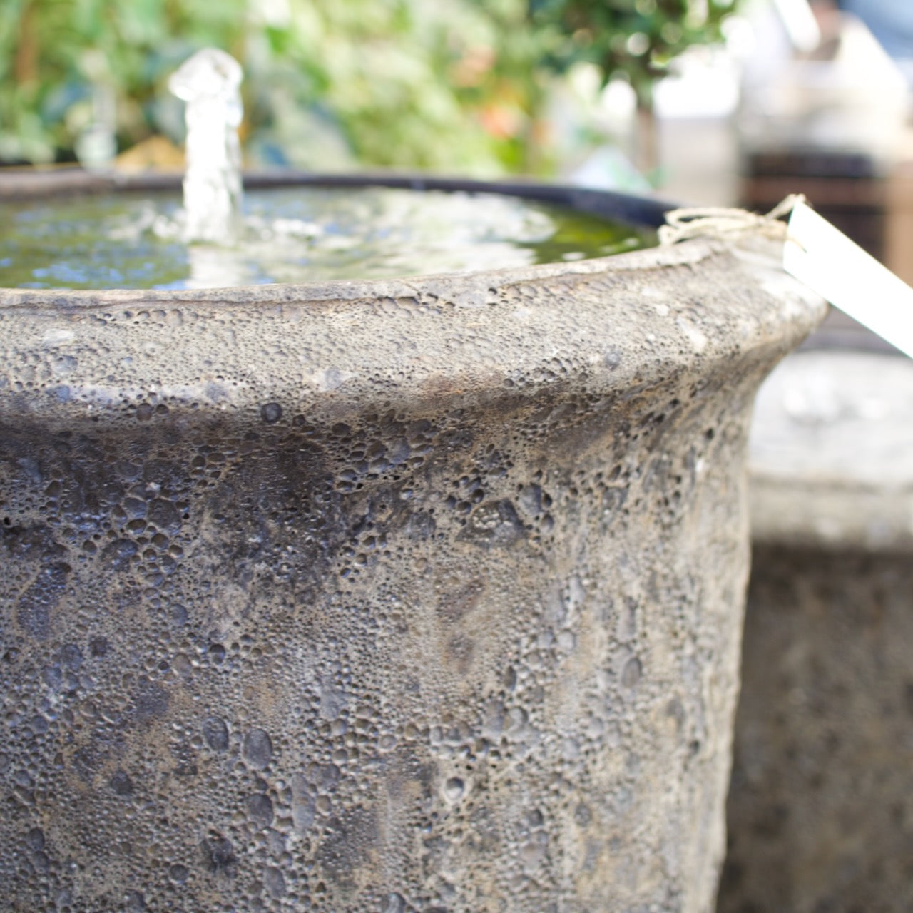 A close-up of a textured, rustic concrete planter or fountain pot with water visible at the top and a small white tag attached near the rim.