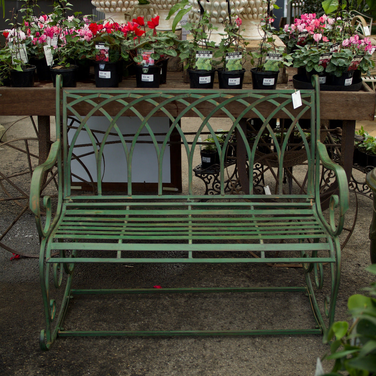 A green metal garden bench with a decorative arched backrest and curved arms, photographed in a nursery or garden center setting with potted flowers on a table behind it.