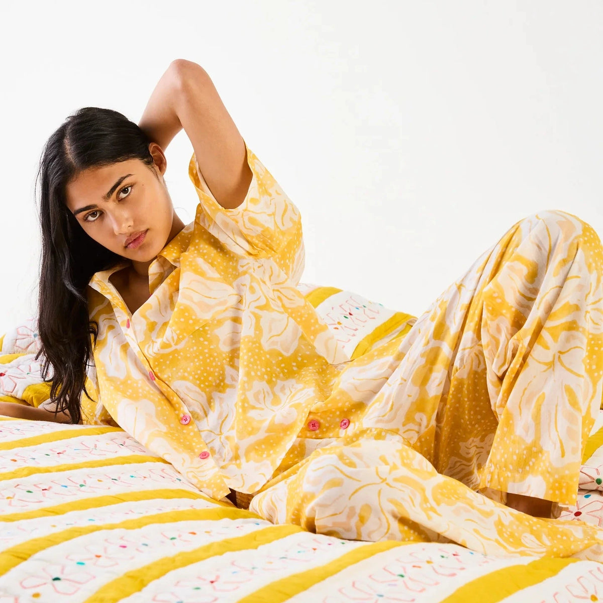 A woman reclining on a bed wearing a yellow and white floral pajama set, with a matching yellow and white striped quilt visible.