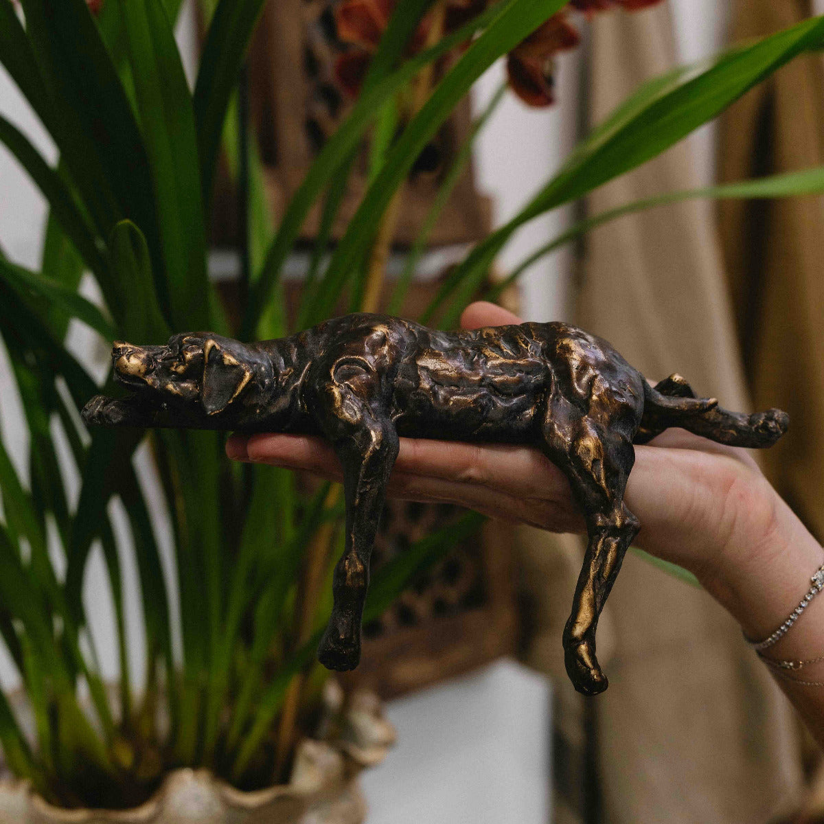 A bronze-finished sculpture of a lying dog being held in a person's hand, with a potted plant and indoor setting blurred in the background.