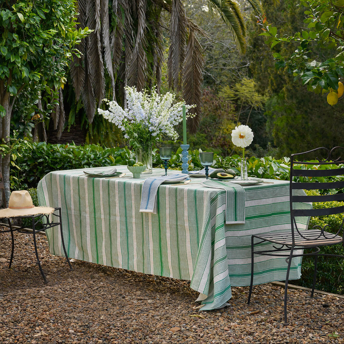 A garden dining setup with a table draped in a green and white striped tablecloth, set with white flowers, blue glassware, and black metal chairs, surrounded by lush greenery and a palm tree.