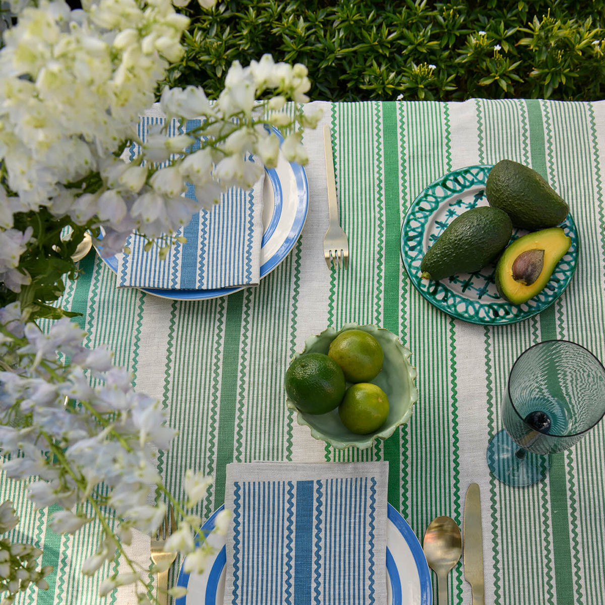 An overhead view of a styled outdoor table setting with a green and white striped tablecloth, blue and white plates, striped napkins, a bowl of limes, and a plate with avocados, with white flowers in the foreground.