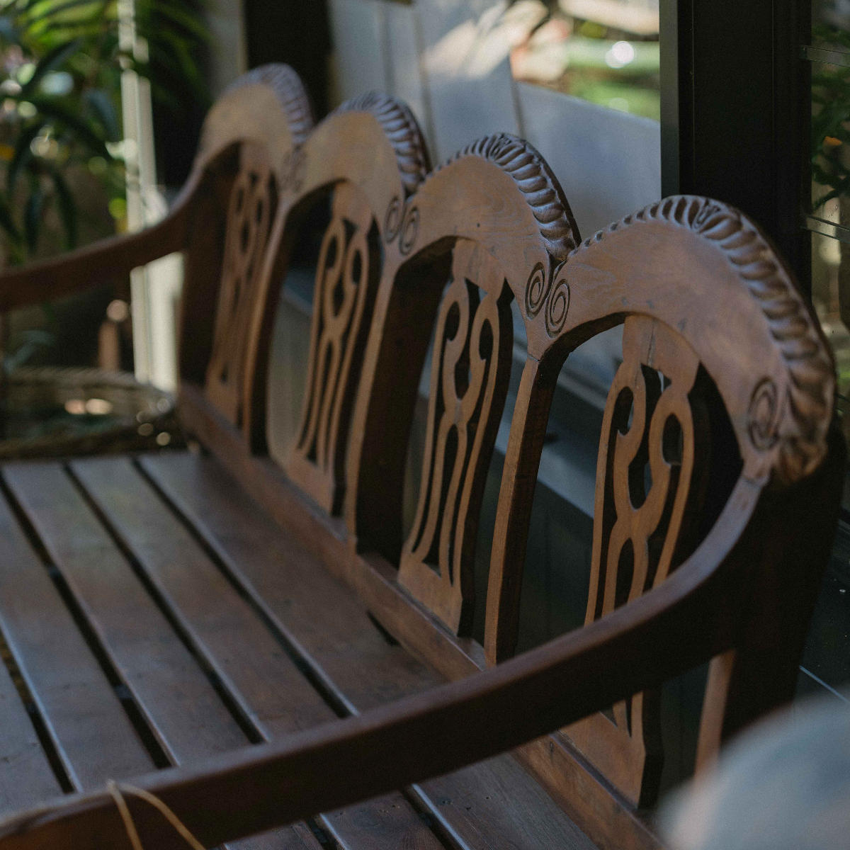 A close-up of a carved wooden bench with ornate backrests and a slatted seat, photographed in a real outdoor porch or garden setting.
