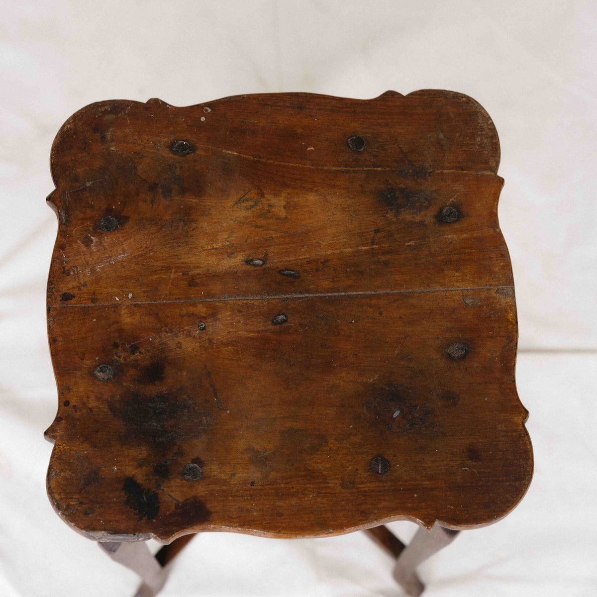 A top-down view of a rustic, square wooden stool or table top with scalloped edges and dark metal hardware, set against a white fabric background.