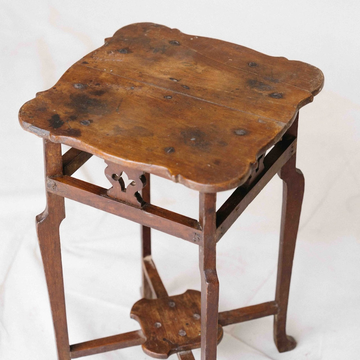 An antique wooden side table with a scalloped top and curved legs, shown on a white fabric background.