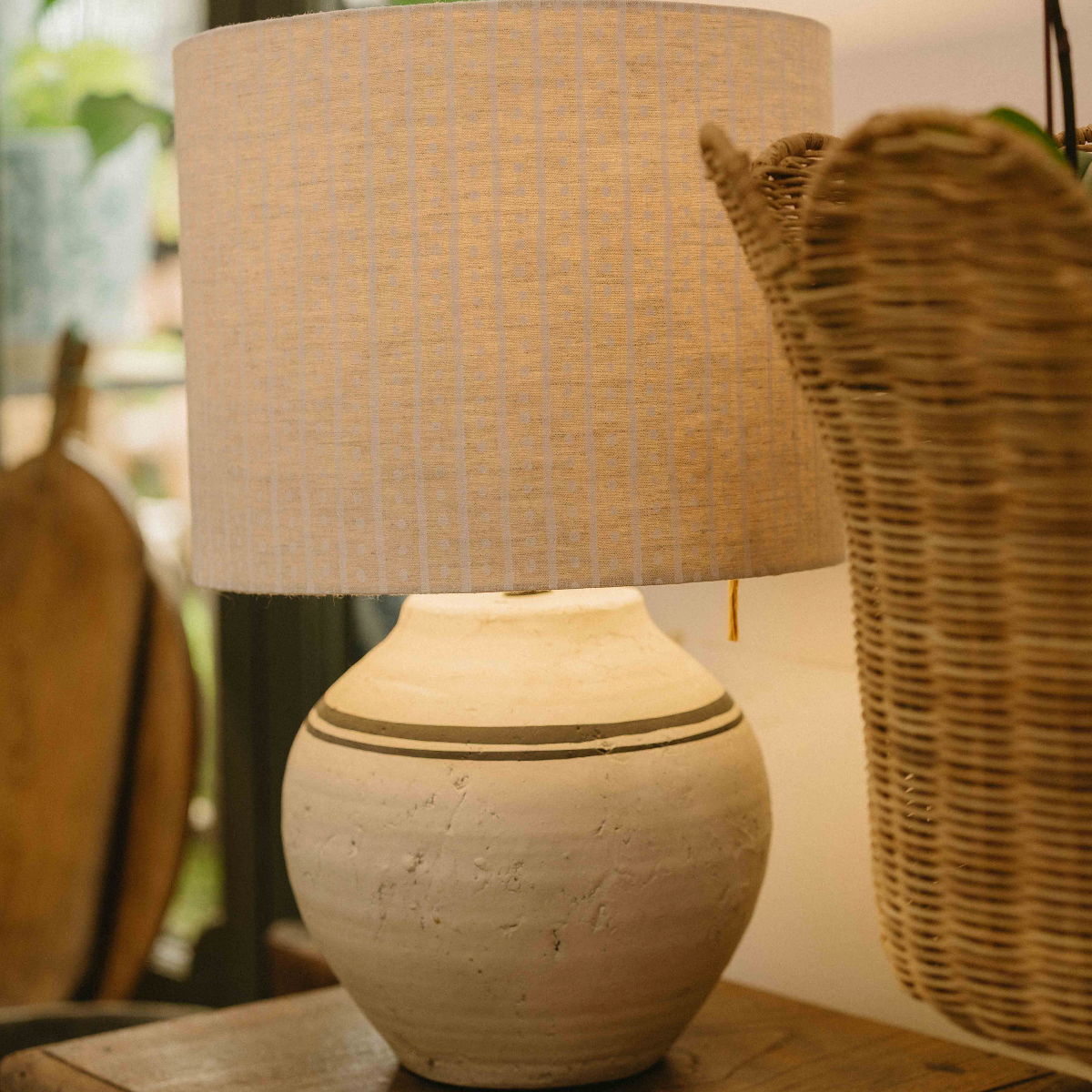 A rustic table lamp with a textured beige linen drum shade and a round, earthy ceramic base with two dark horizontal stripes, set on a wooden table next to a wicker basket.