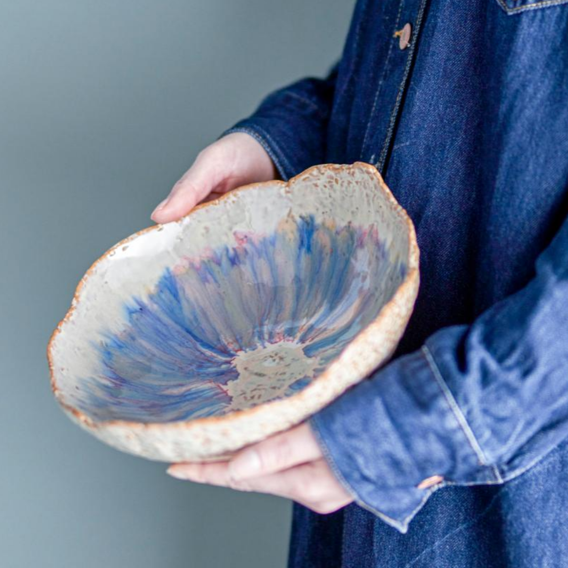 A person in a blue denim jacket holds a handmade ceramic bowl with a rustic, scalloped rim. The bowl features a textured off-white exterior and a vibrant, radial blue, purple, and pink glazed interior.
