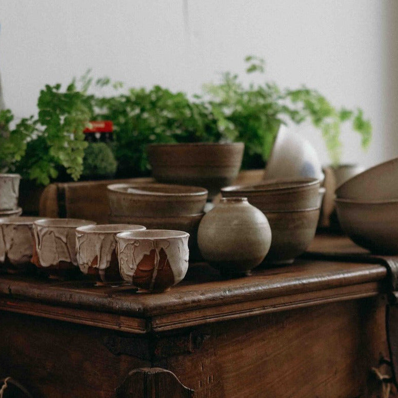 A collection of handmade ceramic pottery, including bowls, cups, and small vases in earthy beige, brown, and gray glazes, displayed on top of a rustic dark wood chest in a styled interior setting.