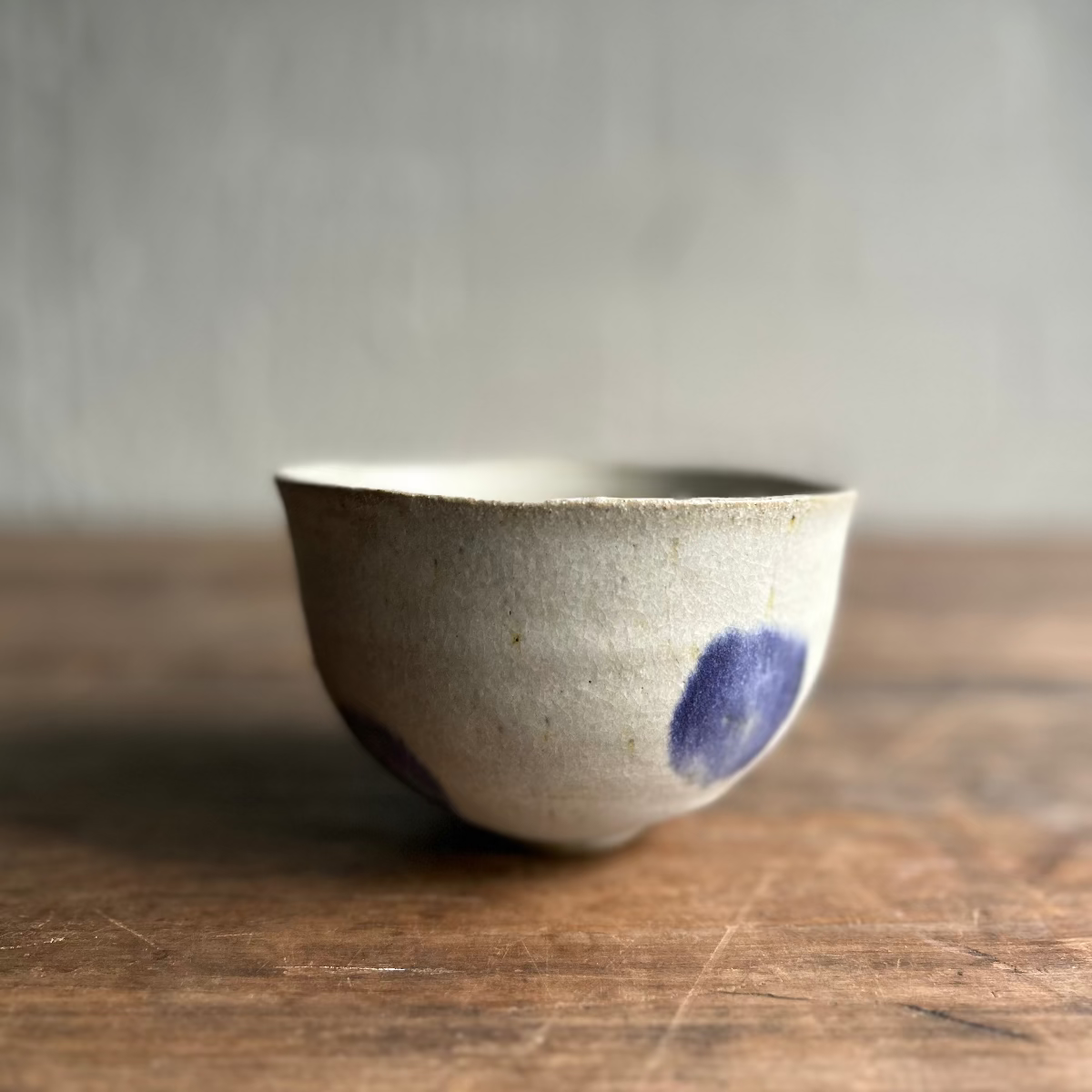 A handmade ceramic bowl with a speckled off-white glaze and blue painted accents, resting on a rustic wooden surface against a neutral gray background.