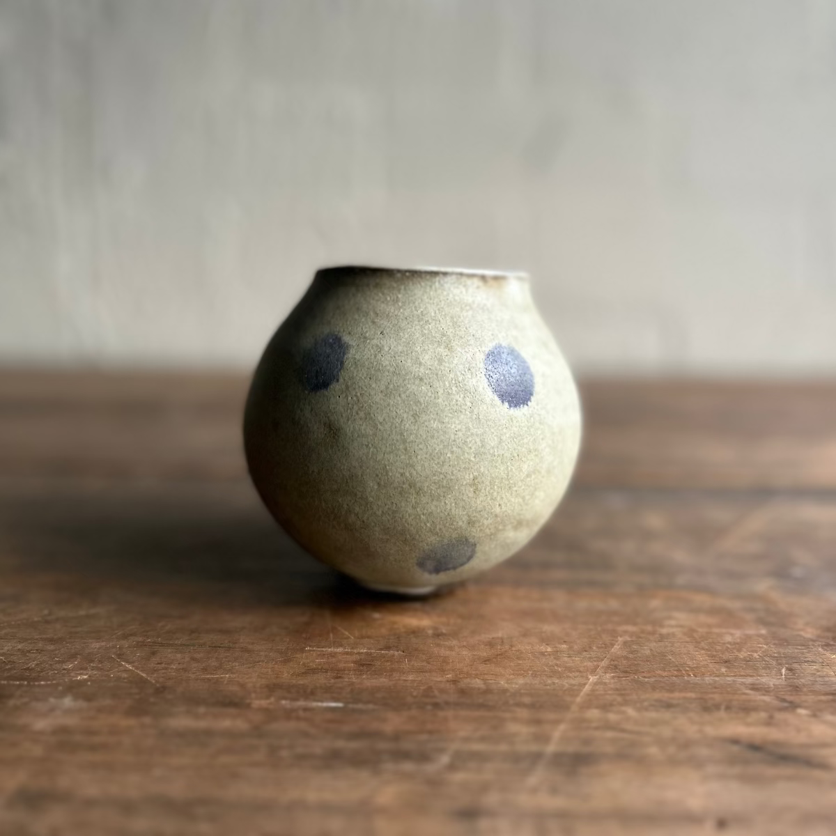 A small handmade ceramic pot with a speckled beige-gray glaze and blue-gray painted dots, sitting on a rustic wooden surface against a textured gray wall.