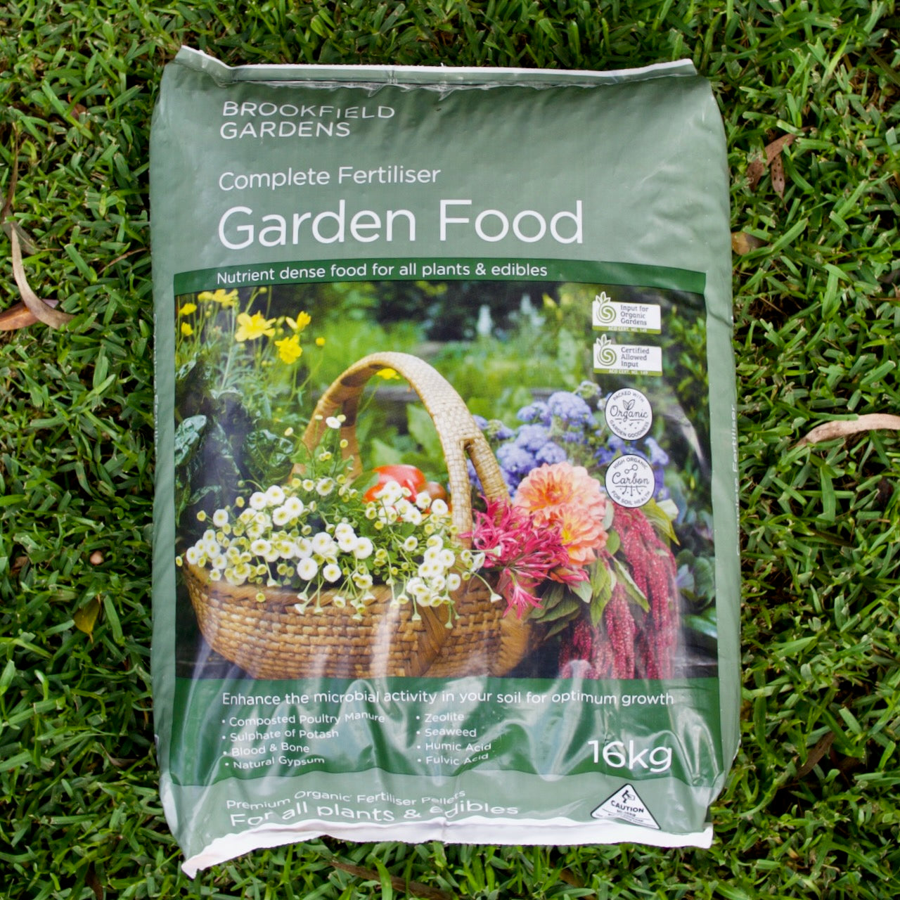 A 16kg bag of Brookfield Gardens Complete Fertiliser Garden Food, placed on green grass. The bag is sage green with a large photo of a wicker basket filled with colorful flowers.