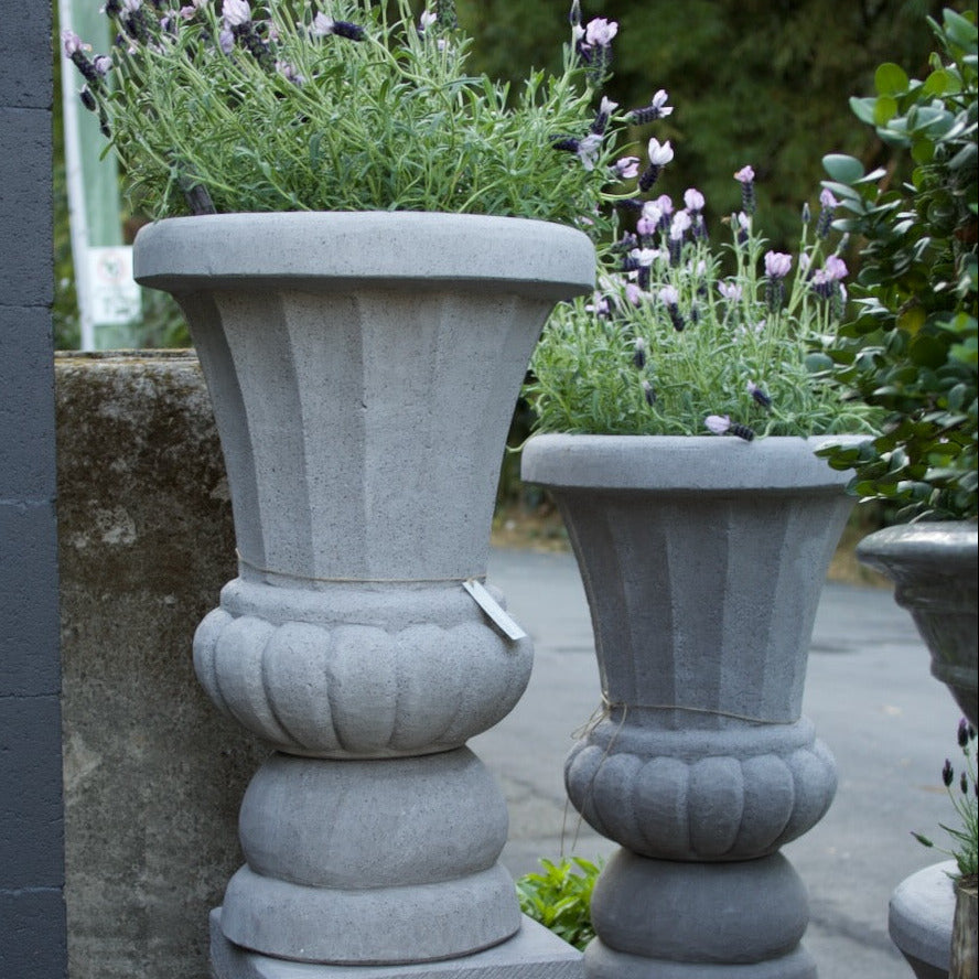Two gray stone urn-shaped planter pots with decorative pedestals, filled with green foliage and light purple flowers, set outdoors next to a dark gray wall.