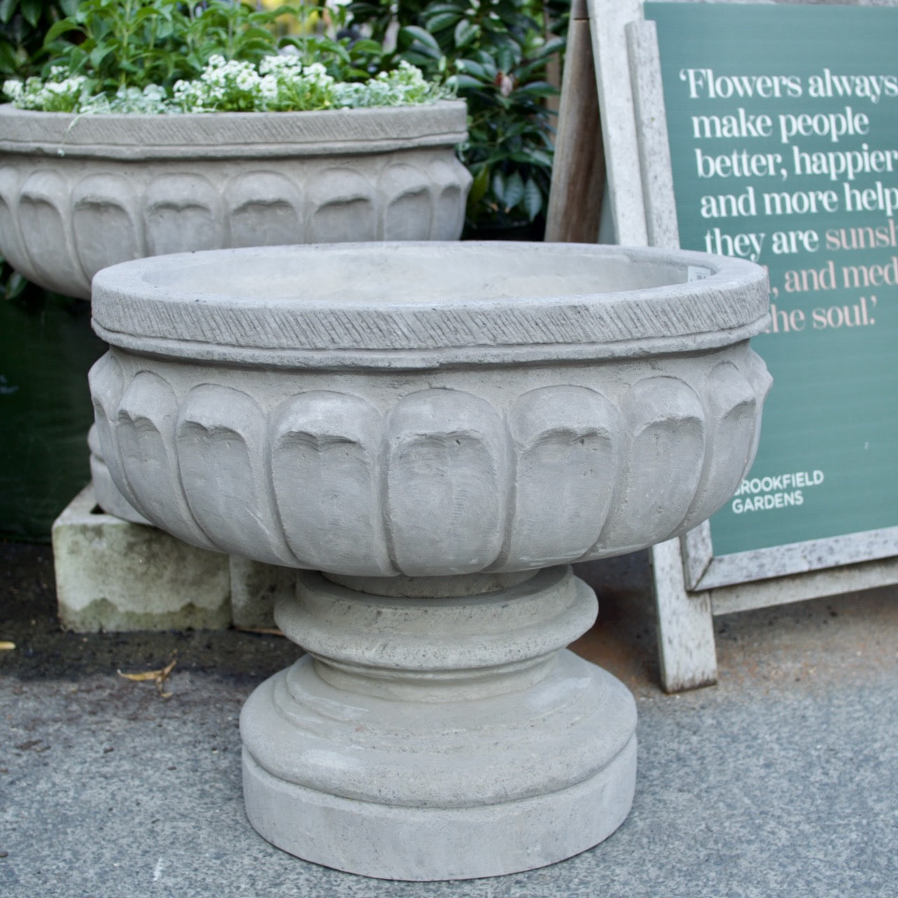 A round concrete planter with a fluted pedestal base and a wide rim, shown outdoors on a paved surface next to other planters and a garden sign.