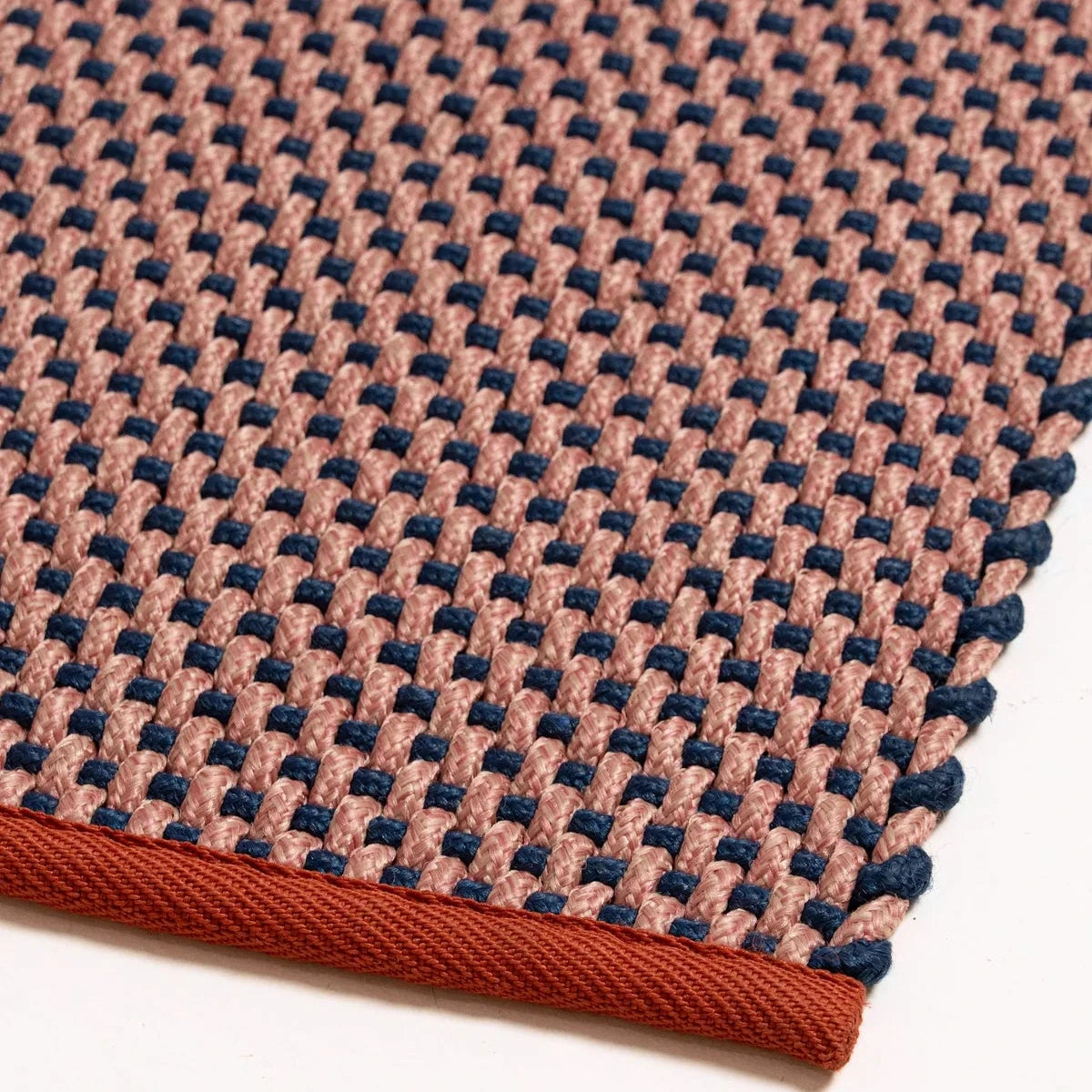 A close-up of a woven textile product, likely a rug or mat, showing a textured pattern of dusty pink and navy blue woven loops with a rust-brown braided edge on a white background.