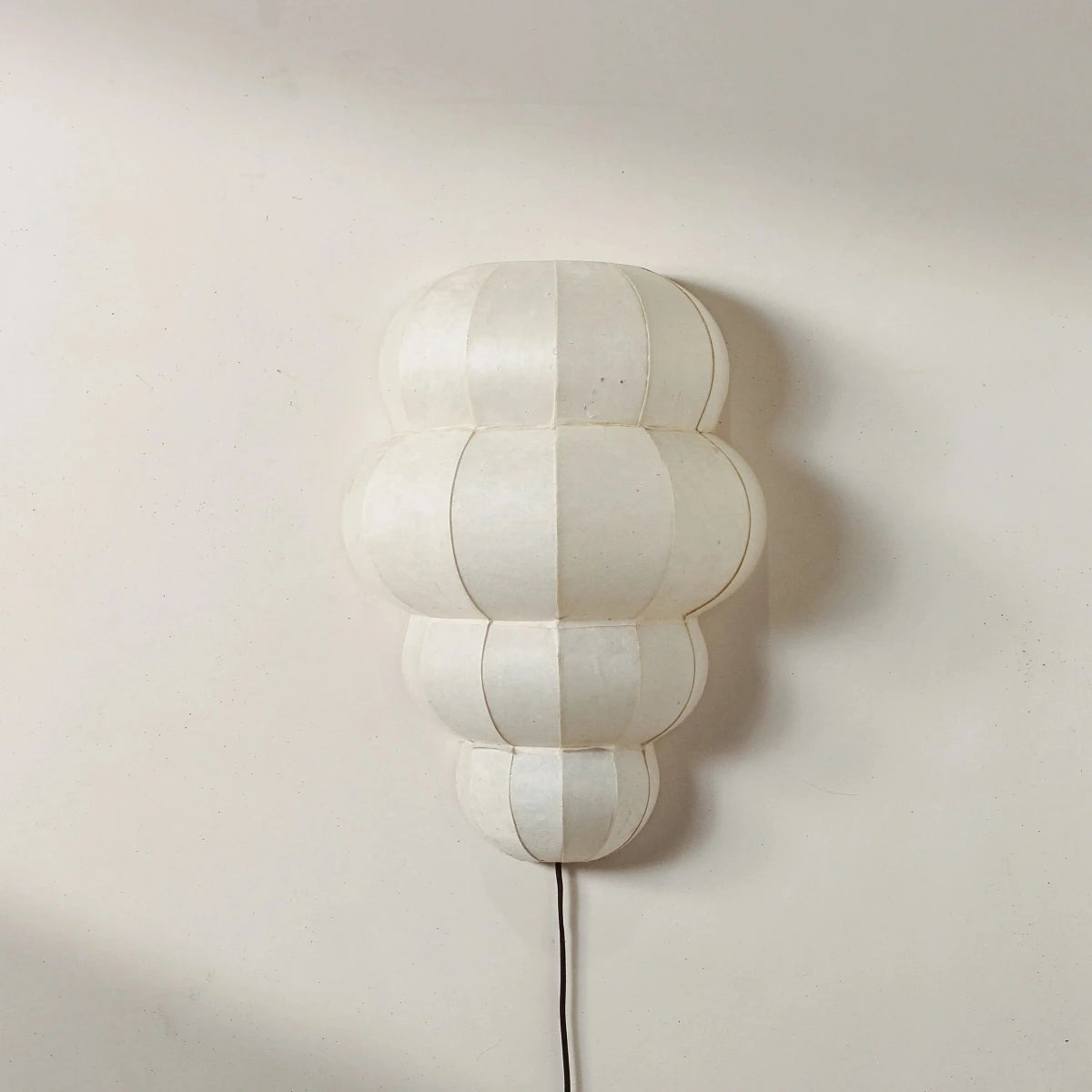 A modern wall-mounted light fixture with a segmented, cloud-like organic shape in an off-white or cream color, mounted on a plain beige wall with a black power cord visible below.